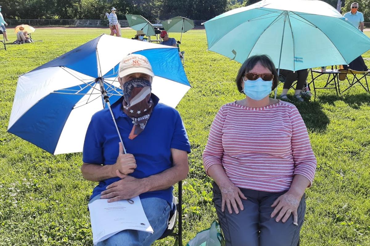 Man and woman wearing masks sitting under umbrellas on sunny day